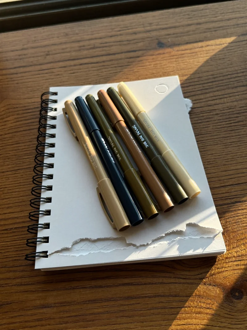 Fineliners og alkoholmarkers i vifte ved siden af blank spiralskitseblok i blødt vinduslys